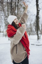 A cheerful young woman smiles as she lifts her hand in a snowy park, dressed warmly with a red