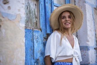 A smiling woman in a straw hat leans against a rustic blue door in Trypiti, Milos Island, Greece.