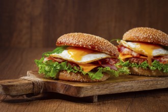 Two juicy burgers sit on a wooden board, each filled with lettuce, bacon, cheese, and fried eggs