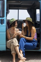 Two lesbian women enjoy leisure time by a camper van, sipping coffee and wearing casual attire.