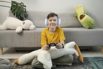 A young boy in a yellow shirt plays video games while sitting cross-legged on a rug. Surrounded by