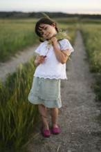A young girl, identifiable by her feminine dress and hairstyle, stands on a dirt path with wheat