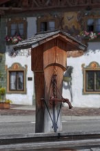 A traditional wooden fountain in front of a Bavarian building with decorative flowers and sunshine,