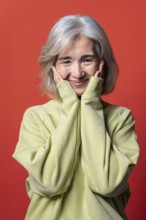 A joyful Asian Chinese senior woman with gray hair and a yellow sweater poses cheerfully against a