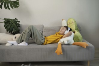 A young boy in a yellow shirt lies comfortably on a grey sofa, wearing headphones and surrounded by