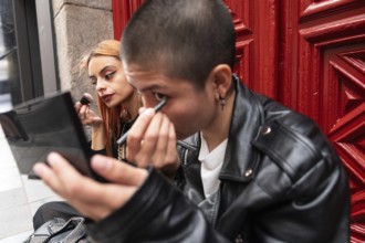 A lesbian couple sits together in front of a red door, each applying makeup with focus and care,