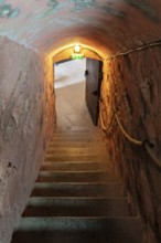 A dimly lit stone tunnel with a staircase and a rope railing leading to an open door area, Le