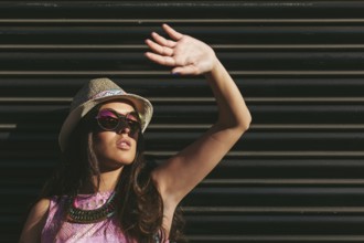 Woman wearing a summer hat and sunglasses raises hand to shield eyes from the sun. The background