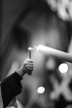 A monochrome image depicting a hand holding a lighter, reaching to ignite a tall candle,