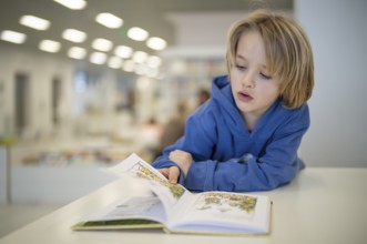 Interior shot, boy, 6 years old, blonde, reading book in the children's section of the city
