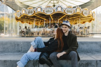 A young couple sits on steps in front of a brightly lit carousel in Brooklyn Bridge Park. They