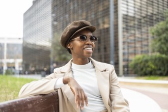 A fashionable black woman in sunglasses and a hat sits on a city bench, exuding confidence and