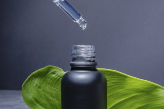 A dropper releasing a clear serum into a matte black bottle, set against a solid grey background