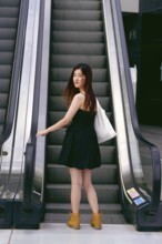 A young asian woman in a black dress and boots stands on an escalator in an urban setting, holding