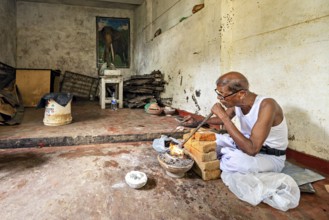 An old man works silently in a workshop with bricks and fire, an elephant poster on the wall, a man