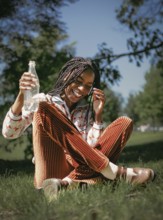 A joyful African American woman with braided hair laughs heartily while sitting on grass and