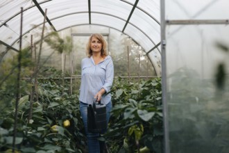 A woman stands in a lush greenhouse, holding a watering can. Surrounded by thriving plants, she
