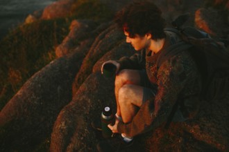 A young male hiker sits on rugged rocks at Cape Spear Lighthouse, watching the sunset. He is