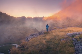 A lone person stands on a grassy ridge, gazing at misty mountains during dusk in summer. Warm light