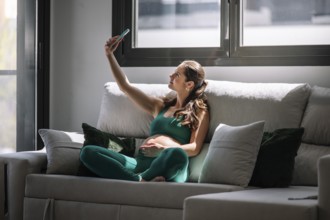 Pregnant woman sitting comfortably on a couch, wearing a yoga outfit, and taking a selfie. She