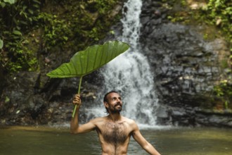 Shirtless man holding a large leaf smiles as he uses it for shade while standing in a water pool in