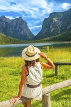 Woman wearing straw hat admiring lake dobbiaco with the dolomites in the background on a sunny