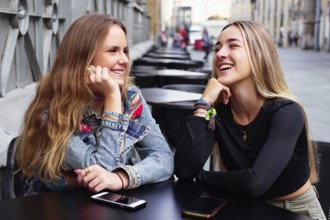 Two teenage girls smile and chat outdoors, with cell phones on the table, enjoying each other's