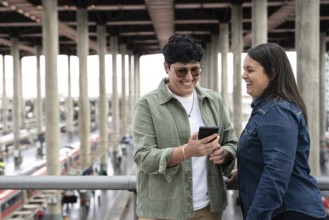 A joyful moment between a lesbian curvy couple at a train station, sharing smiles and a phone,