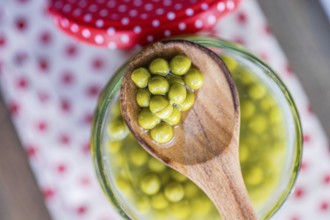 A wooden spoon holds green homemade organic peas over a glass jar, set on a white cloth with red