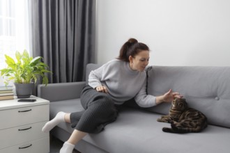 Woman gently pets her cat on a gray sofa in a cozy living room. A potted plant and books add to the