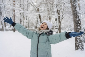 A cheerful child in a blue jacket and white hat enjoys a snowy day in a winter forest, embracing