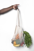 A hand holds a reusable cotton mesh bag filled with fresh vegetables and fruits, showcasing