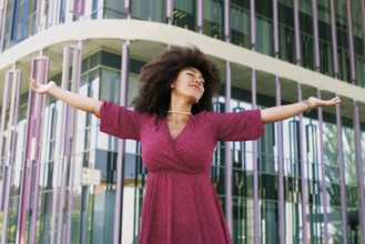 Young Arab woman with afro hair, wearing a pink dress, joyfully extends her arms in front of a