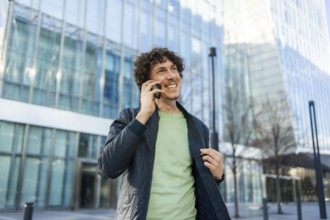 A businessman stands outside a sleek, modern glass building, smiling as he talks on his smartphone,