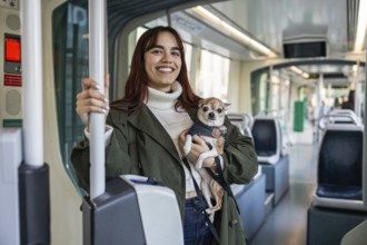 A woman smiles while holding her small chihuahua on a tram. The chihuahua looks calmly at the