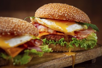 Two juicy burgers sit on a wooden board, each filled with lettuce, bacon, cheese, and fried eggs