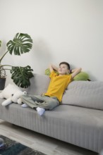 A young boy in a yellow shirt relaxes on a gray couch, leaning against green avocado plush toy and
