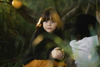 A young child with thoughtful expression sits in a forest setting. Warm sunlight filters through