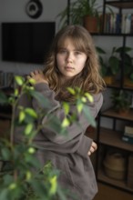 A teenage girl stands thoughtfully in a cozy home, surrounded by houseplants and a bookshelf. Her