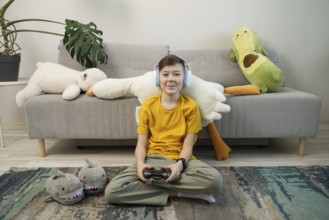 A cheerful boy in a yellow shirt and headphones plays video games, seated in a cozy room with plush