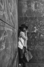 A woman wearing a stylish hat and casual attire leans against a textured, urban wall, exuding