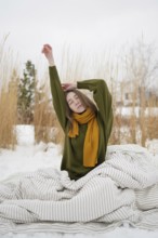 A woman with a serene expression stretches in a snowy park, wearing a green sweater and yellow