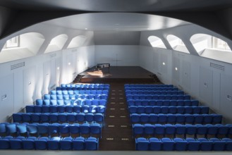 A spacious auditorium with rows of vibrant blue seats facing a stage. Natural light streams through