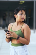 A teenage girl holds a smartphone while standing outdoors. Dressed in a green top and light pants,