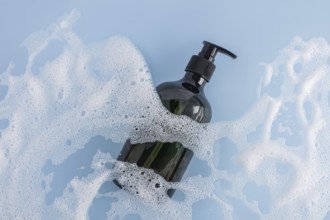 A sleek black shower gel bottle lies amidst generous white foam on a soothing blue background.