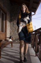 A woman stands on a rustic porch holding her purebred papillon dog. Natural light illuminates the