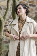 A fashion woman in a beige trench coat and patterned dress stands gracefully outdoors. The stone
