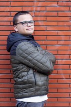 A young man with an intellectual disability stands confidently with arms crossed against a brick