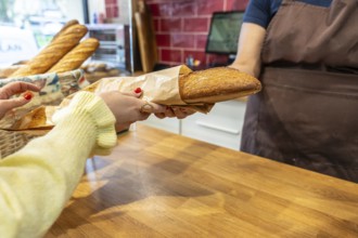 Customer hands collecting a freshly baked baguette wrapped in paper from a baker behind a wooden