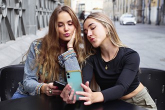 Two teenage girls sit together, using their cell phones and sharing a fun moment. They are
